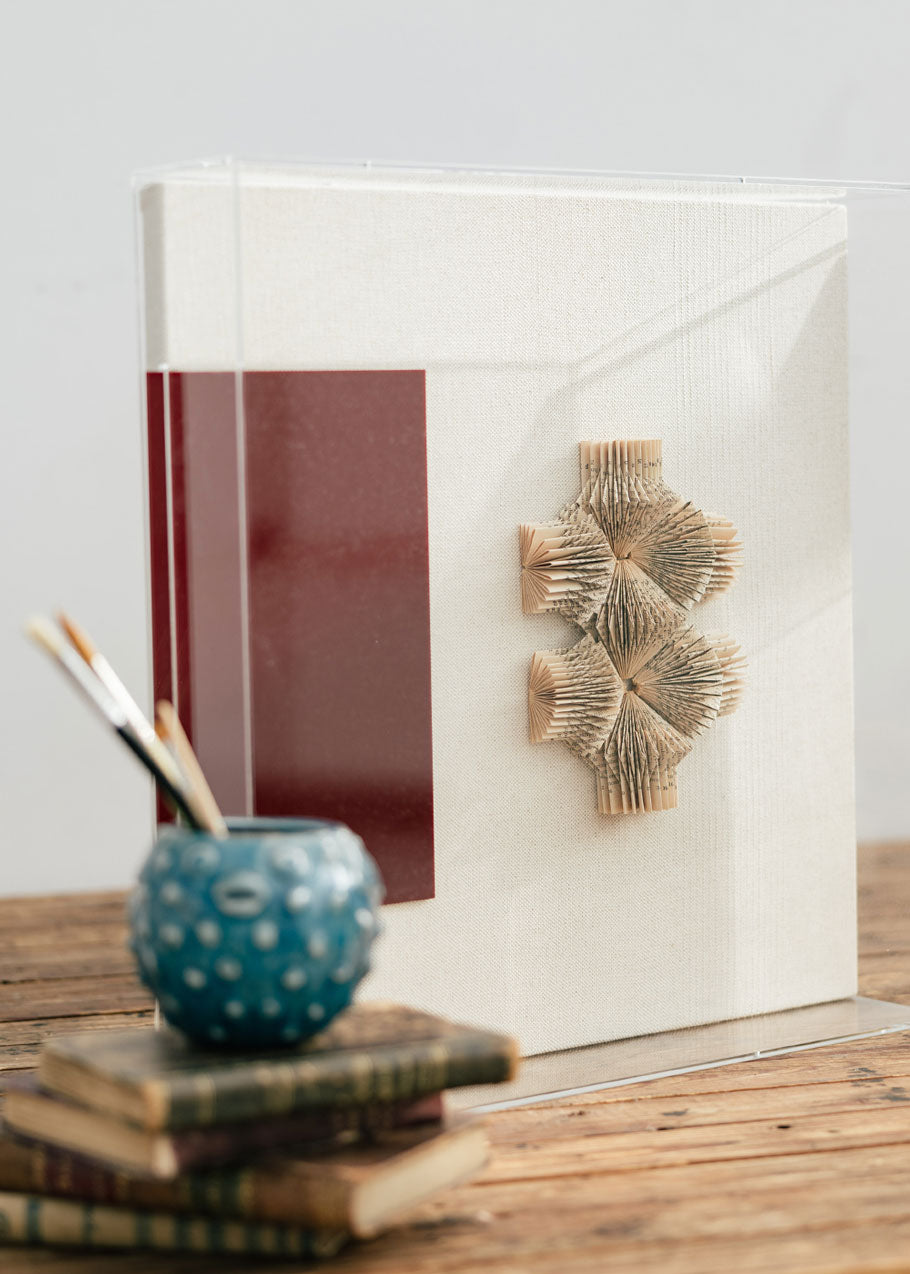 Escultura de pared forrada en lino rústico con caja de metacrilato transparente. Está formada por una serie de libros plegados y formas geométricas en metacrilato