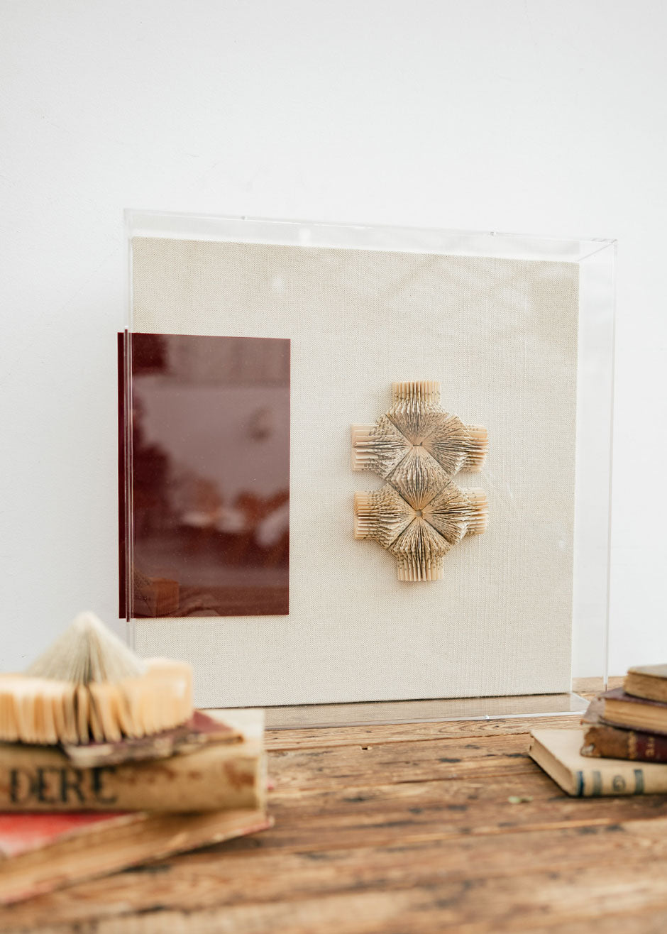 Escultura de pared forrada en lino rústico con caja de metacrilato transparente. Está formada por una serie de libros plegados y formas geométricas en metacrilato