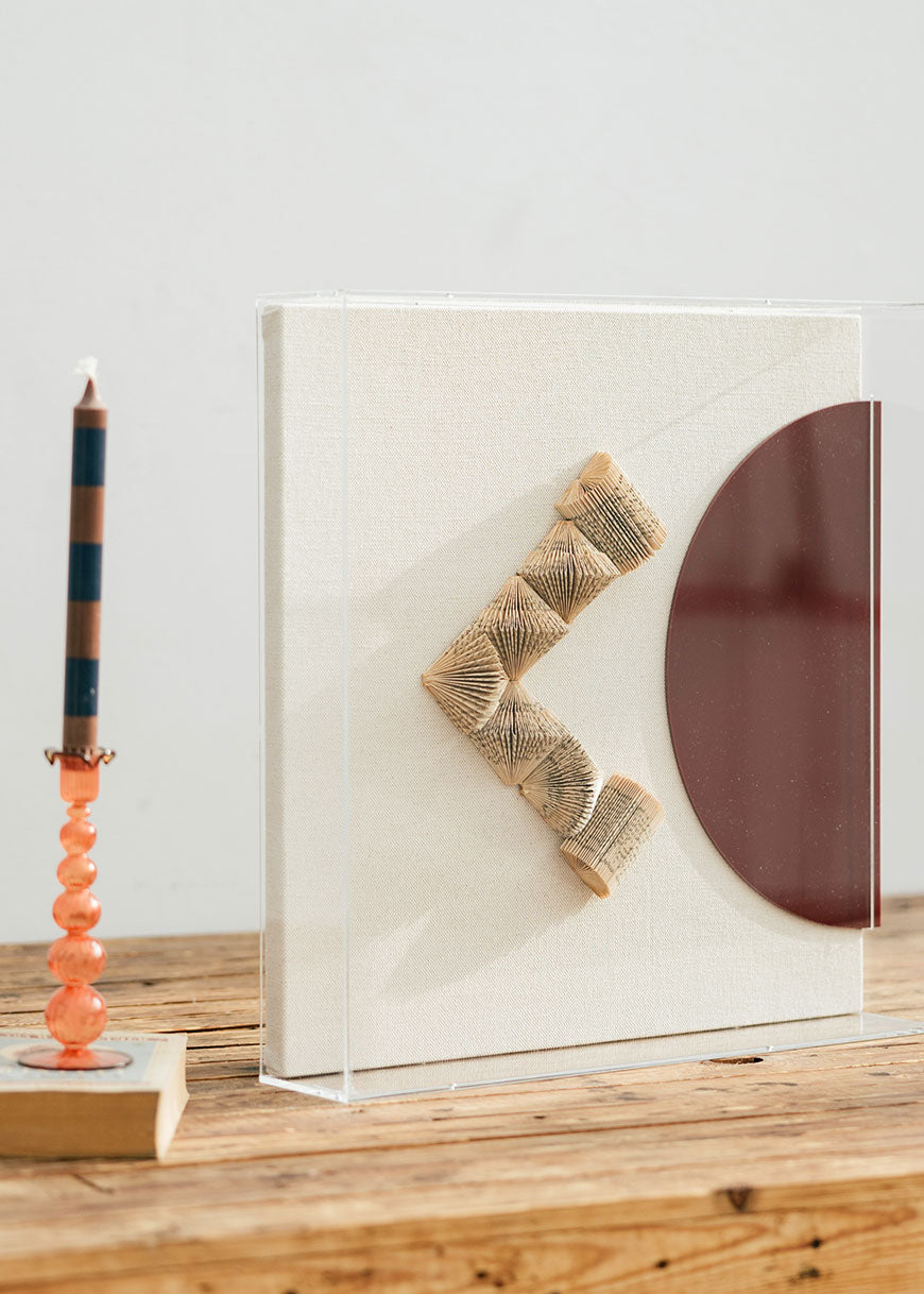 Escultura de pared forrada en lino rústico con caja de metacrilato transparente. Está formada por una serie de libros plegados y formas geométricas en metacrilato.