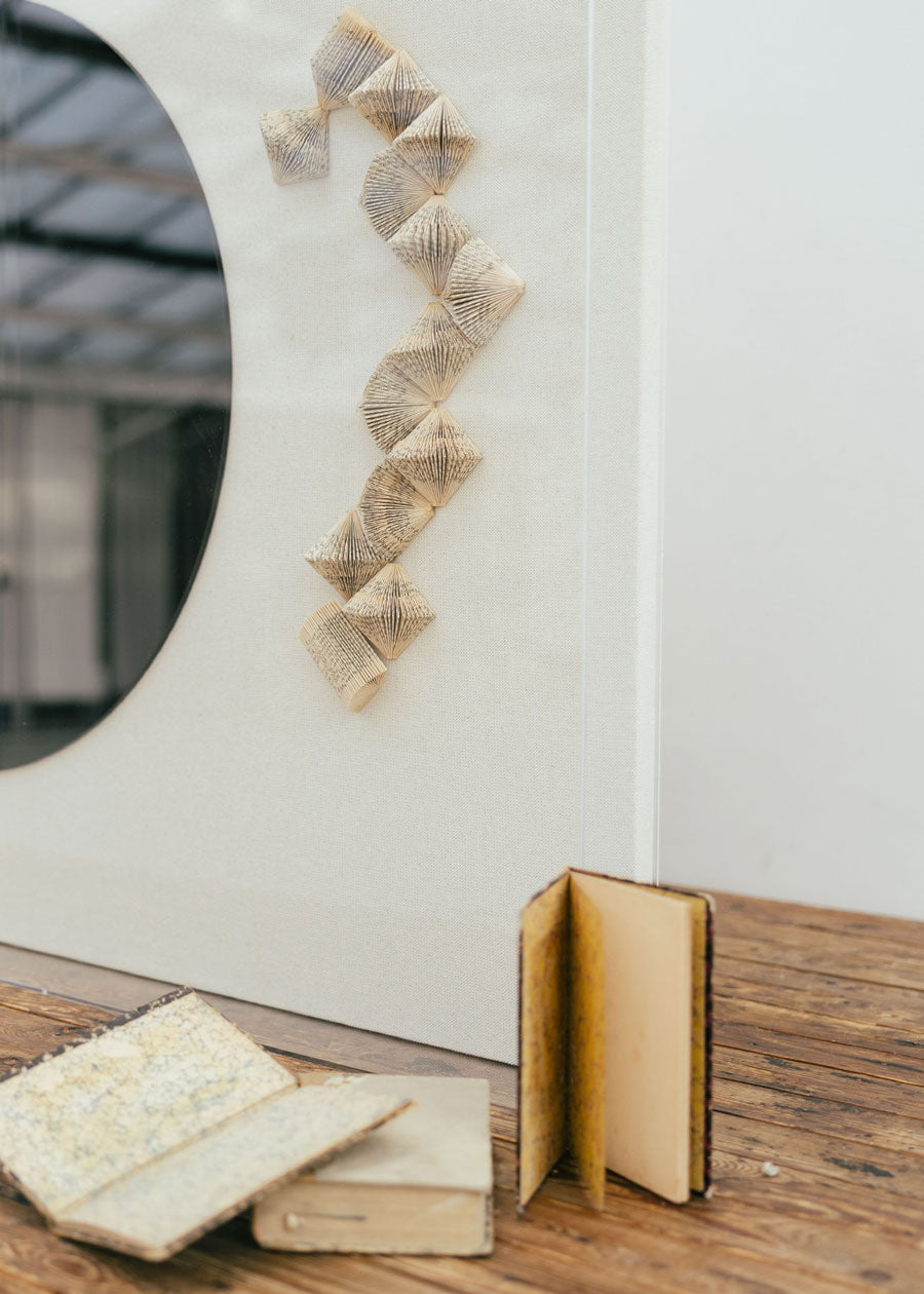 Escultura de pared en dos piezas forradas en lino rústico con cajas de metacrilato transparente. Está formada por una serie de libros plegados y formas geométricas en metacrilato.