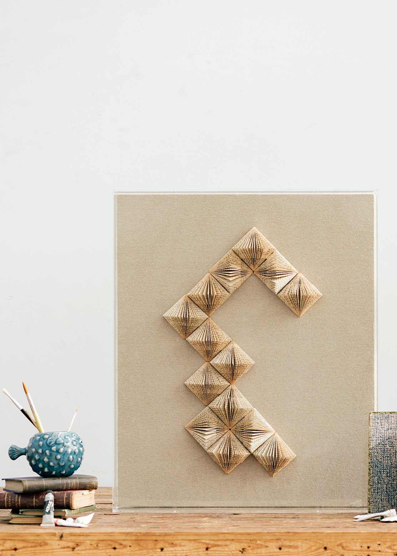 Escultura de pared forrada en lino rústico con caja de metacrilato transparente y serie de libros plegados.
