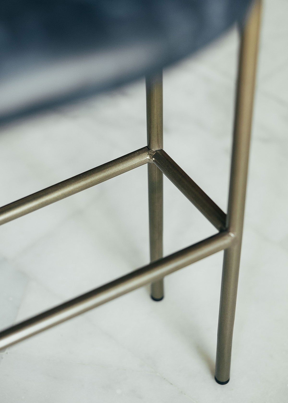 Taburete tapizado en tela de piel azul con estructura de metal acabado en dorado. Un taburete alto con respaldo, reposabrazos y reposapiés incorporado.