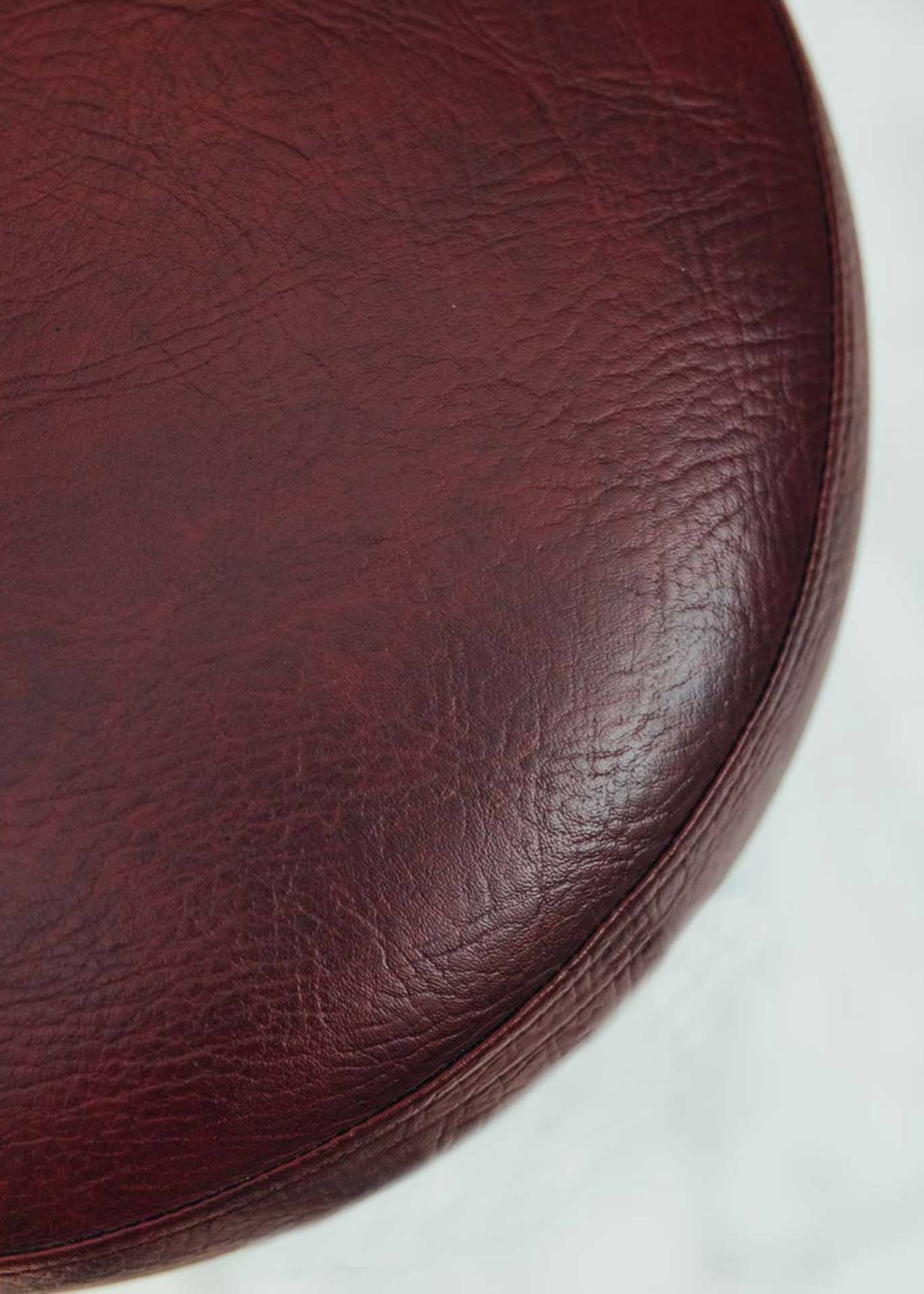 Taburete tapizado con estructura de hierro pavonado compuesta por tubos y base circular. Un taburete alto con reposapiés y asiento tapizado en tela de piel granate.