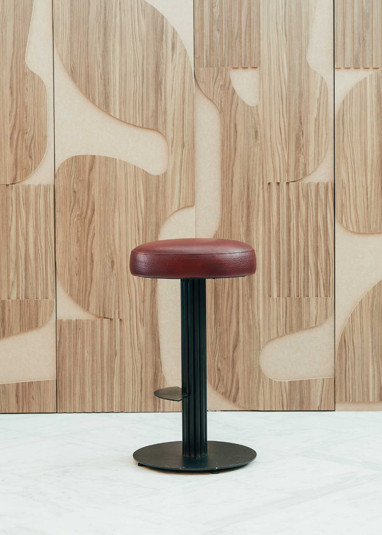 Taburete tapizado con estructura de hierro pavonado compuesta por tubos y base circular. Un taburete alto con reposapiés y asiento tapizado en tela de piel granate.