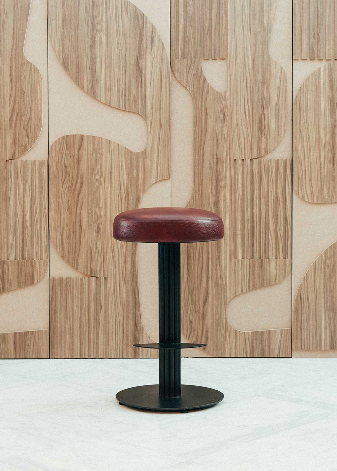 Taburete tapizado con estructura de hierro pavonado compuesta por tubos y base circular. Un taburete alto con reposapiés y asiento tapizado en tela de piel granate.