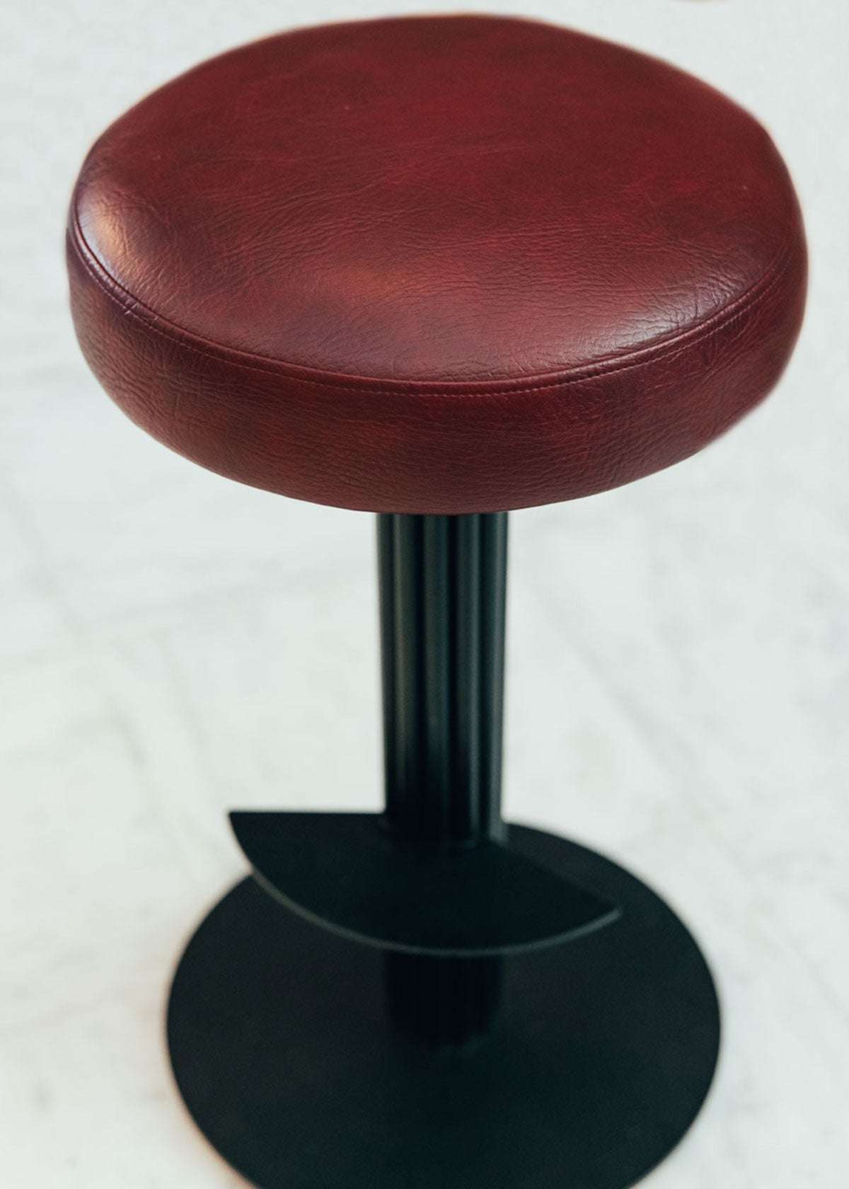 Taburete tapizado con estructura de hierro pavonado compuesta por tubos y base circular. Un taburete alto con reposapiés y asiento tapizado en tela de piel granate.
