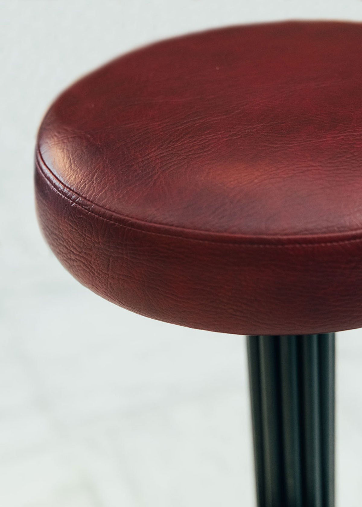 Taburete tapizado con estructura de hierro pavonado compuesta por tubos y base circular. Un taburete alto con reposapiés y asiento tapizado en tela de piel granate.