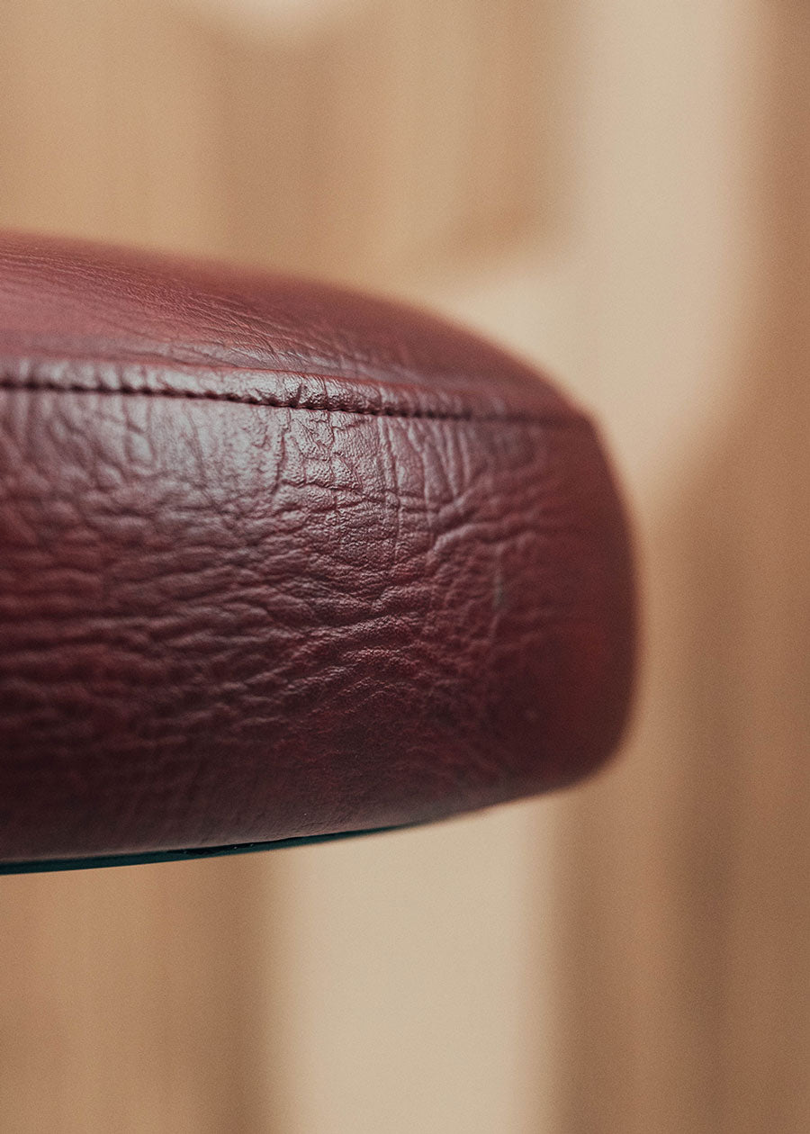 Taburete tapizado con estructura de hierro pavonado compuesta por tubos y base circular. Un taburete alto con reposapiés y asiento tapizado en tela de piel granate.