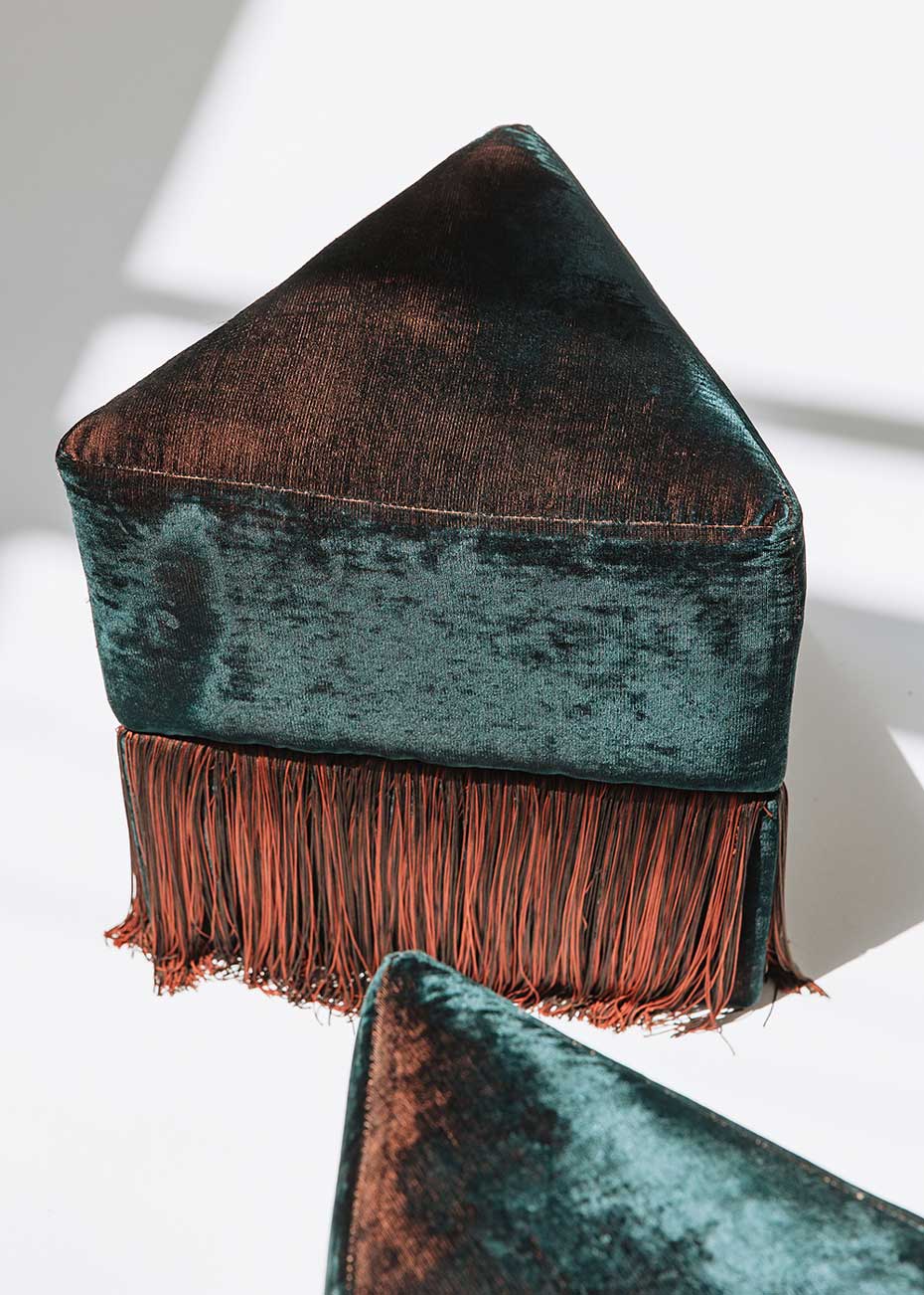 Puf triangular tapizado en terciopelo azul con detalle de flecos en la parte inferior.