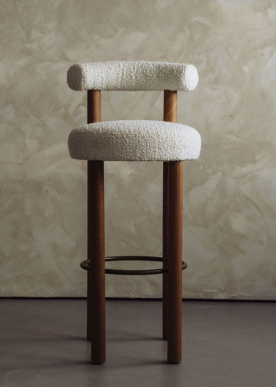 Taburete tapizado en tela beige con estructura de madera de haya. Un taburete alto con respaldo semicircular y reposapiés con acabado de latón incorporado.