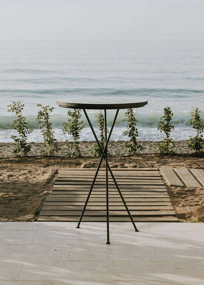 Mesa alta de exterior circular con estructura de tres pies cruzados de hierro acabado en negro.