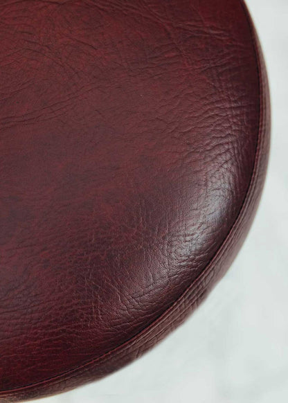 Taburete tapizado con estructura de hierro pavonado compuesta por tubos y base circular. Un taburete alto con reposapiés y asiento tapizado en tela de piel granate.