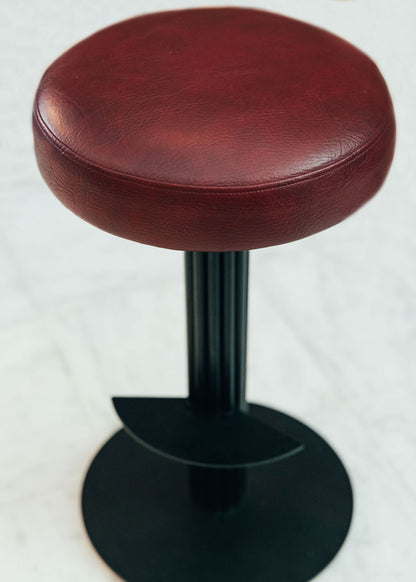 Taburete tapizado con estructura de hierro pavonado compuesta por tubos y base circular. Un taburete alto con reposapiés y asiento tapizado en tela de piel granate.