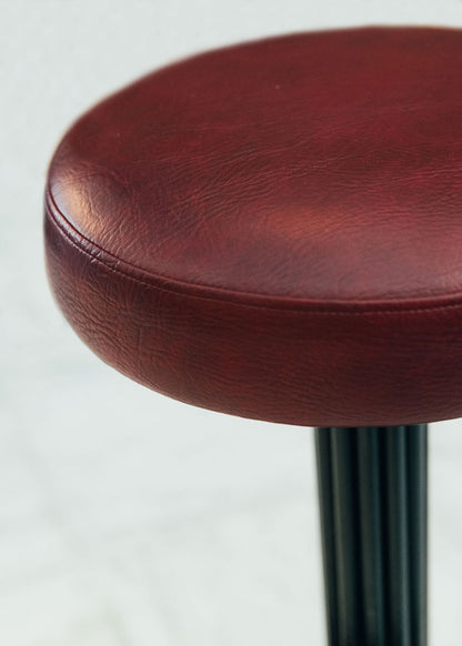 Taburete tapizado con estructura de hierro pavonado compuesta por tubos y base circular. Un taburete alto con reposapiés y asiento tapizado en tela de piel granate.