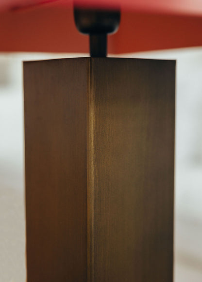 Lámpara de mesa escultórica con base y pantalla metálicas. Base en latón envejecido y pantalla lacada en dos tonos: rojo Burdeos en la cara exterior y color maquillaje en la interior.