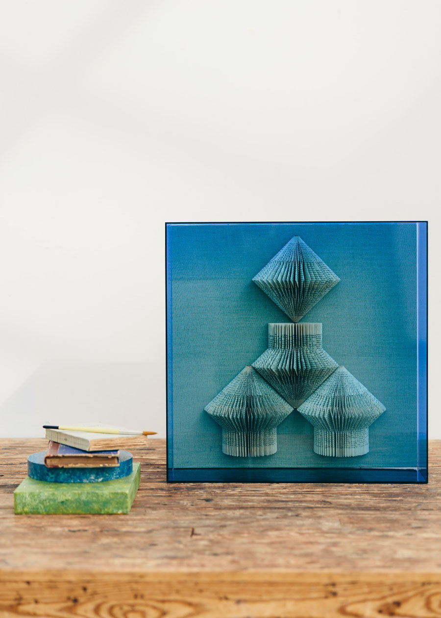 Escultura de pared forrada en lino rústico con caja de metacrilato azul y serie de libros plegados.