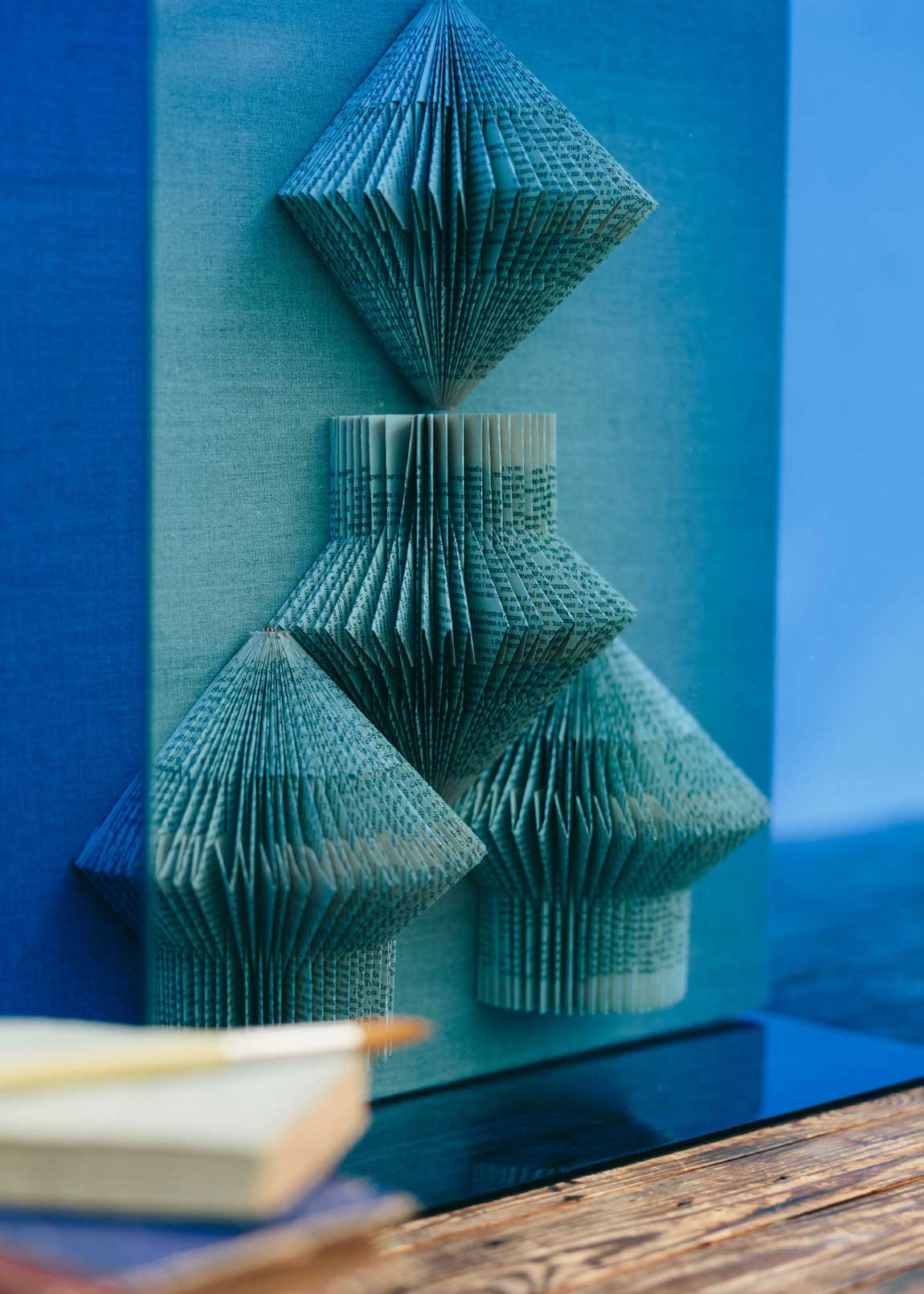 Escultura de pared forrada en lino rústico con caja de metacrilato azul y serie de libros plegados.