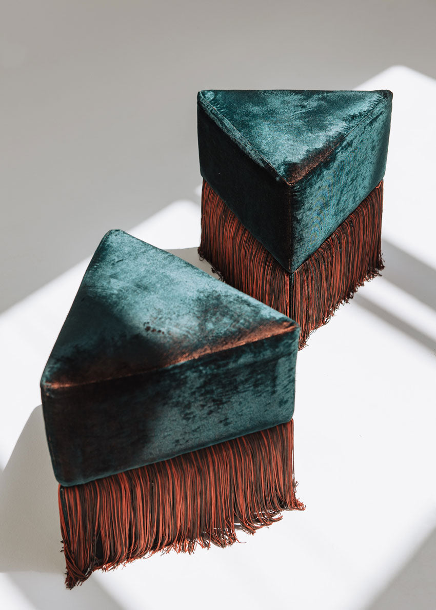 Puf triangular tapizado en terciopelo azul con detalle de flecos en la parte inferior.