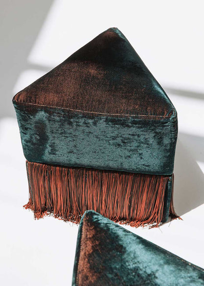 Puf triangular tapizado en terciopelo azul con detalle de flecos en la parte inferior.