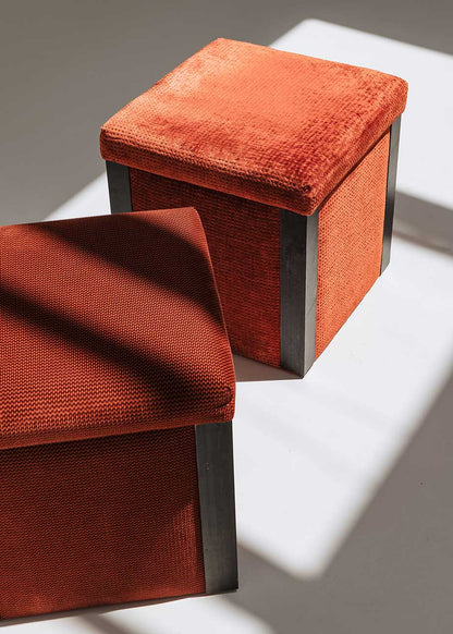 Puf cuadrado tapizado en terciopelo rojo con remates laterales de acero inoxidable.
