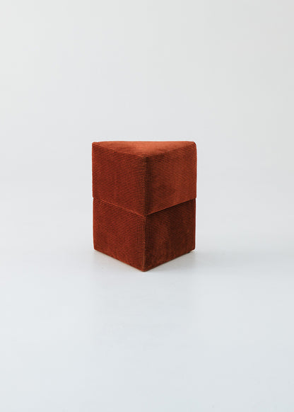 Puf triangular tapizado en tejido en tonos rojos, morados y burdeos.