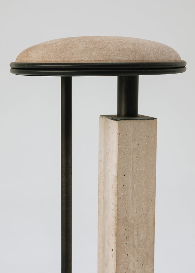 Taburete tapizado con estructura de hierro con detalle de piedra travertino. Un taburete alto con reposapiés y asiento tapizado en tela de terciopelo beige.