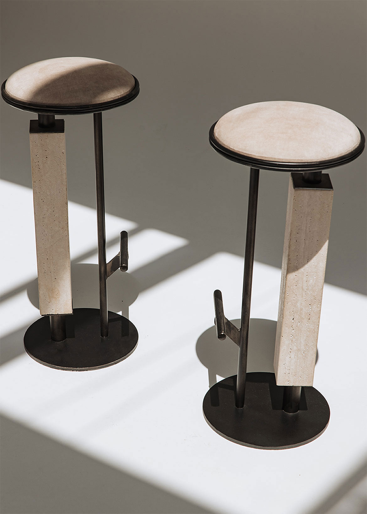 Taburete tapizado con estructura de hierro con detalle de piedra travertino. Un taburete alto con reposapiés y asiento tapizado en tela de terciopelo beige.