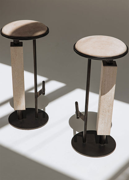 Taburete tapizado con estructura de hierro con detalle de piedra travertino. Un taburete alto con reposapiés y asiento tapizado en tela de terciopelo beige.