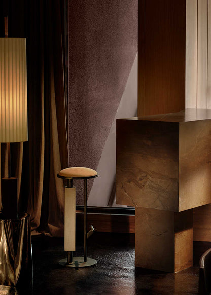 Taburete tapizado con estructura de hierro con detalle de piedra travertino. Un taburete alto con reposapiés y asiento tapizado en tela de terciopelo beige.