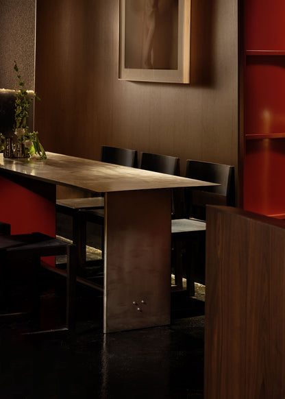 Mesa de comedor rectangular con estructura metálica de acero inoxidable cepillado para patas y sobre. Una de las patas va lacada en rojo.