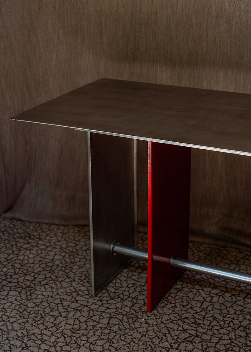 Mesa de comedor rectangular con estructura metálica de acero inoxidable cepillado para patas y sobre. Una de las patas va lacada en rojo.