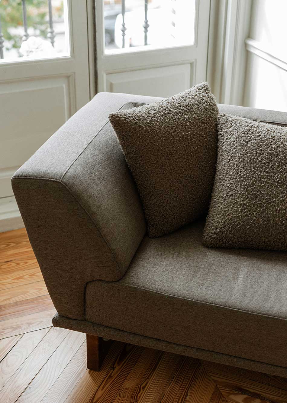 Diván con estructura y patas de madera tapizado con tela gris. Incluye almohadones con relleno en fibra.