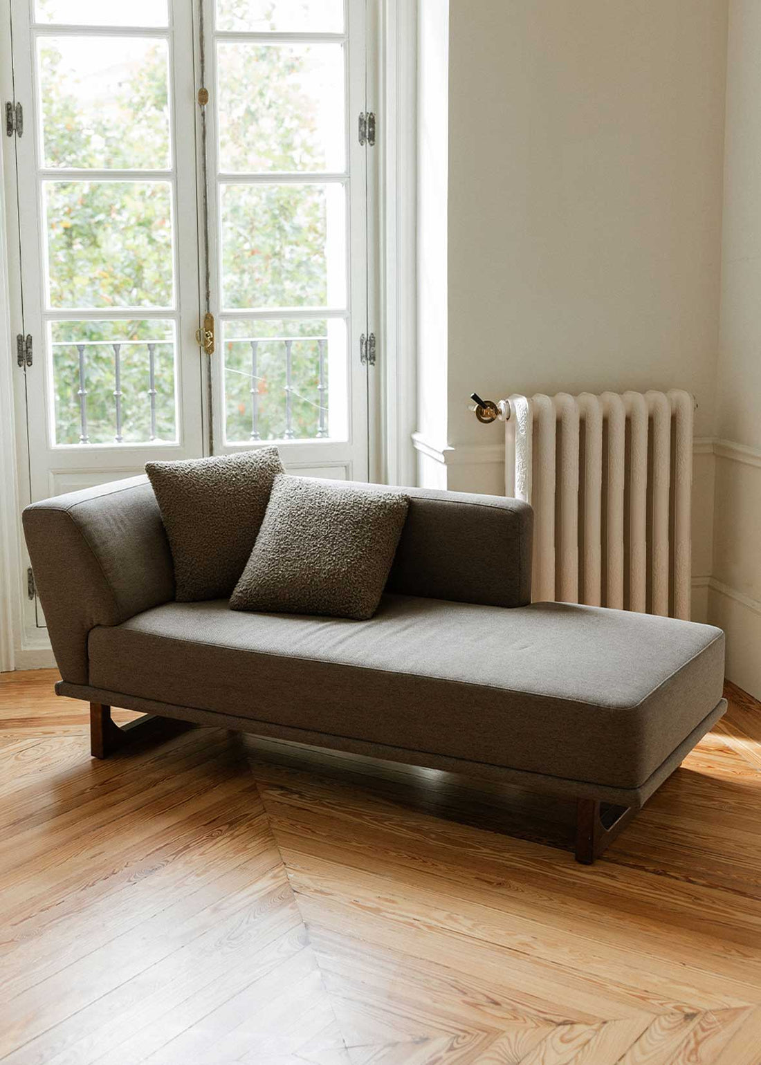 Diván con estructura y patas de madera tapizado con tela gris. Incluye almohadones con relleno en fibra.