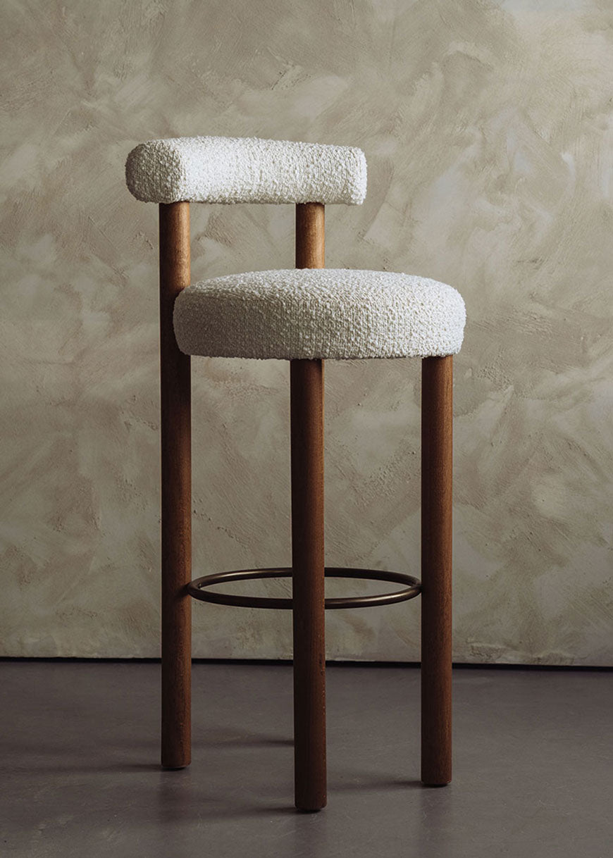 Taburete tapizado en tela beige con estructura de madera de haya. Un taburete alto con respaldo semicircular y reposapiés con acabado de latón incorporado.