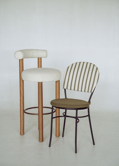 Taburete tapizado en tela beige con estructura de madera de haya. Un taburete alto con respaldo semicircular y reposapiés con acabado de latón incorporado.