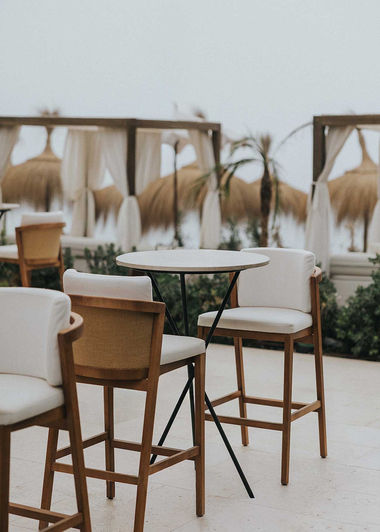 Mesa alta de exterior circular con estructura de tres pies cruzados de hierro forjado acabado en negro y sobre de mármol crema marfil. 