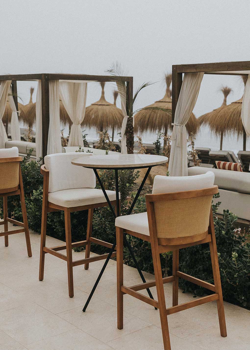 Mesa alta de exterior circular con estructura de tres pies cruzados de hierro forjado acabado en negro y sobre de mármol crema marfil. 