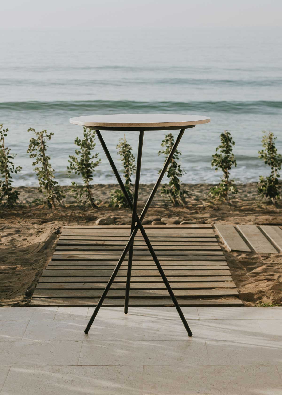 Mesa alta de exterior circular con estructura de tres pies cruzados de hierro forjado acabado en negro y sobre de mármol crema marfil. 