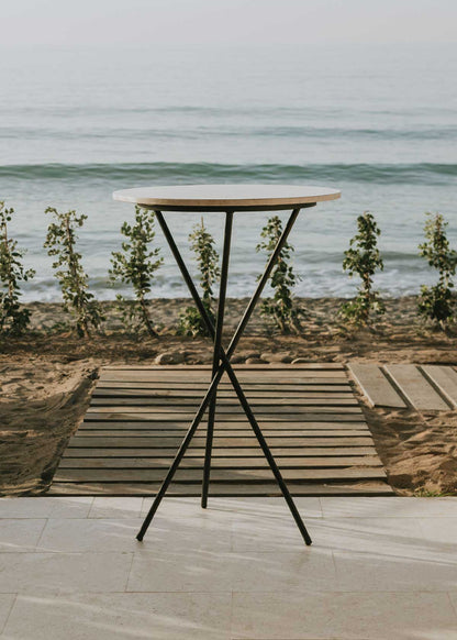 Mesa alta de exterior circular con estructura de tres pies cruzados de hierro forjado acabado en negro y sobre de mármol crema marfil. 