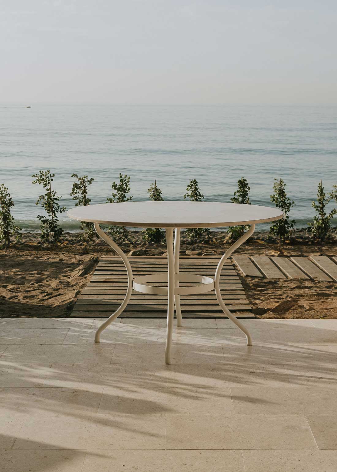 Mesa de exterior circular con estructura de hierro forjado acabado en blanco y sobre de mármol crema marfil.