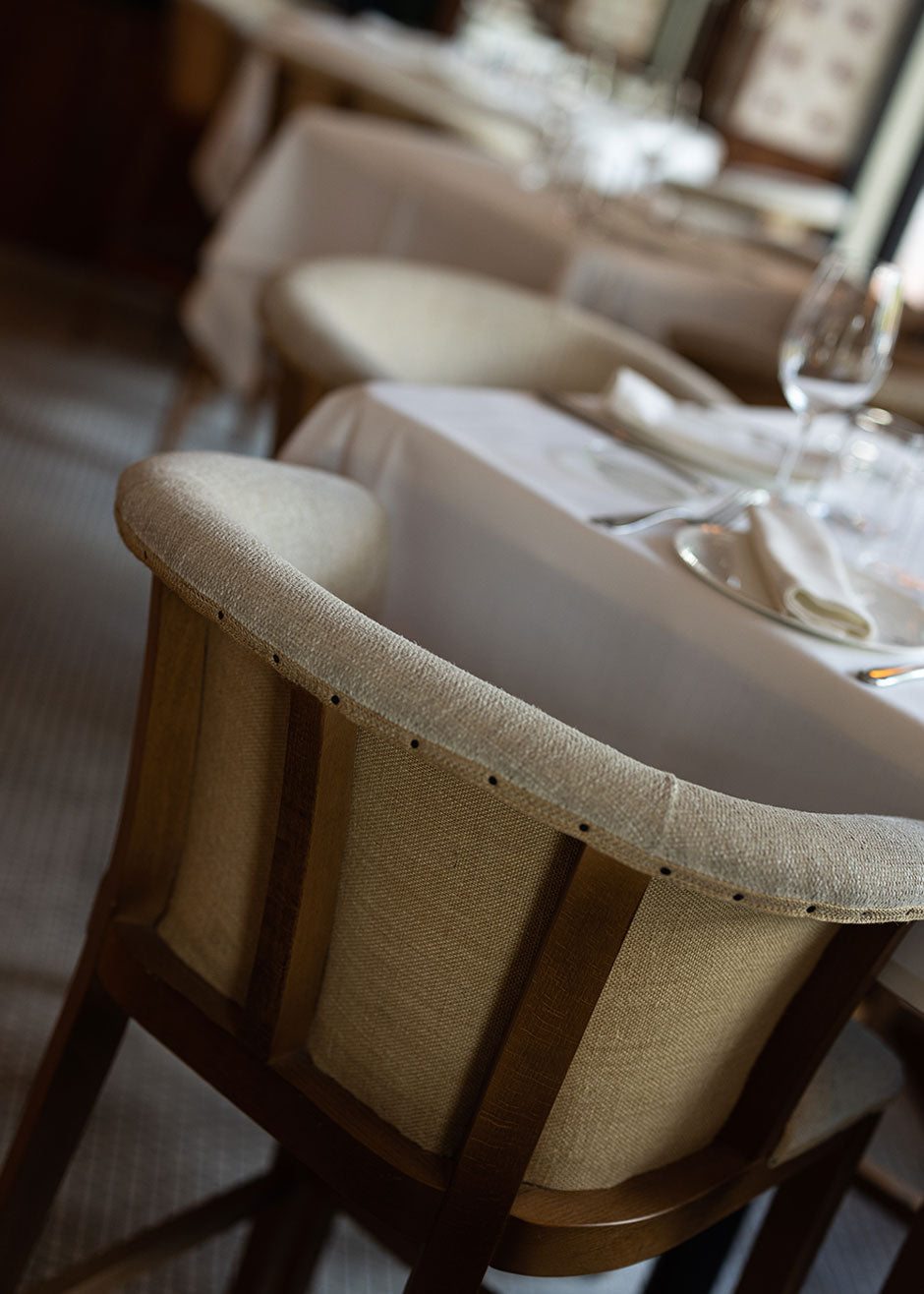 Taburete con estructura de madera de haya, tapizado en tela beige y acabado con un ribete de yute con remaches.